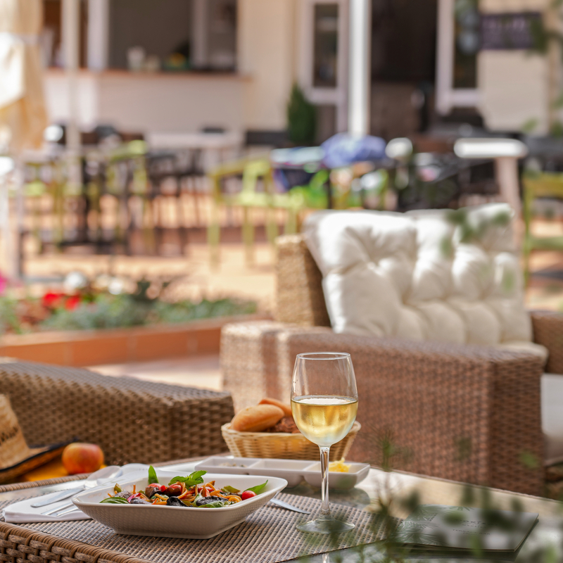 Tisch mit Essen und Weinglas auf einer sonnigen Terrasse im Hotelgarten.