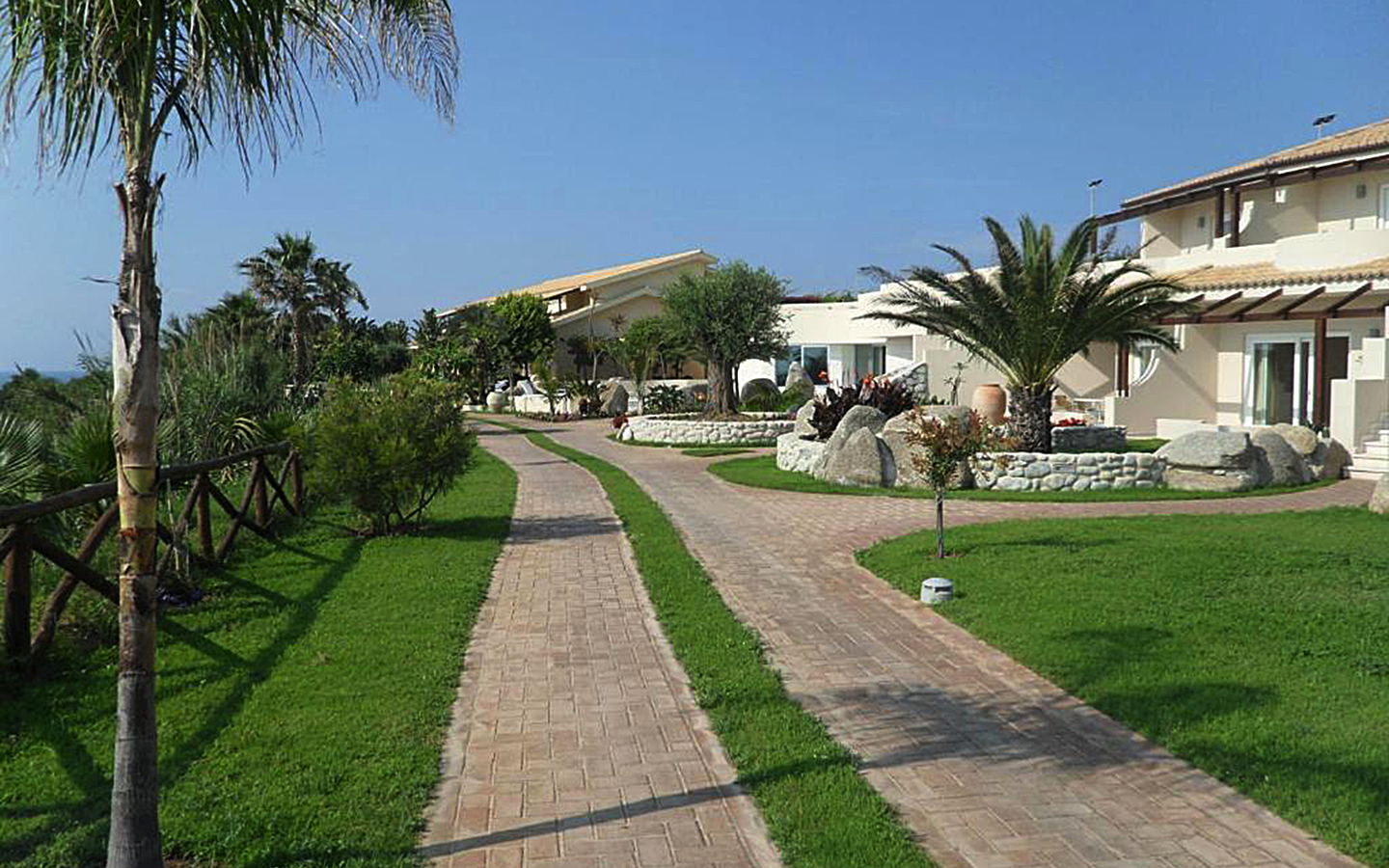 Gepflegter Gartenweg zum Hotelgebäude, umgeben von Palmen und Sträuchern.