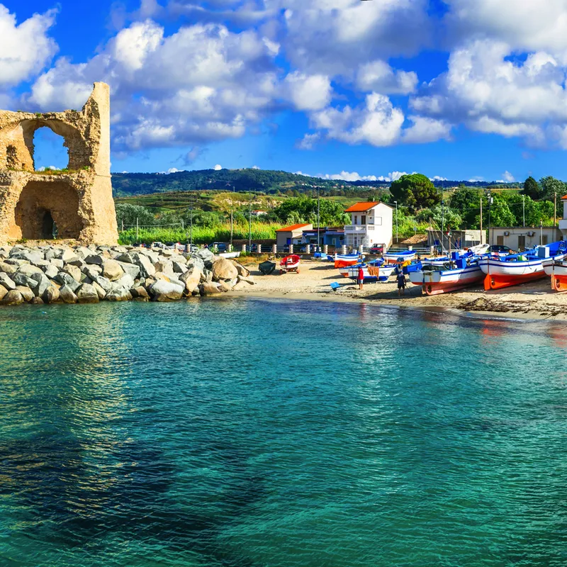 Mittelmeerbucht mit Booten, altem Turm und Häusern bei sonnigem Wetter.
