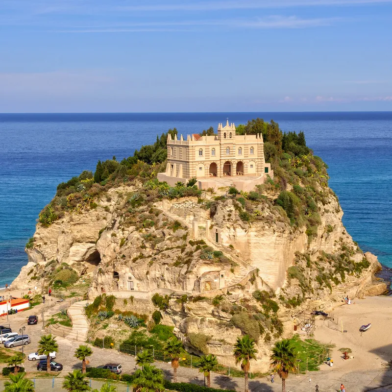 Kloster auf einem Felsen an der Küste mit Strand und blauem Meer im Hintergrund.