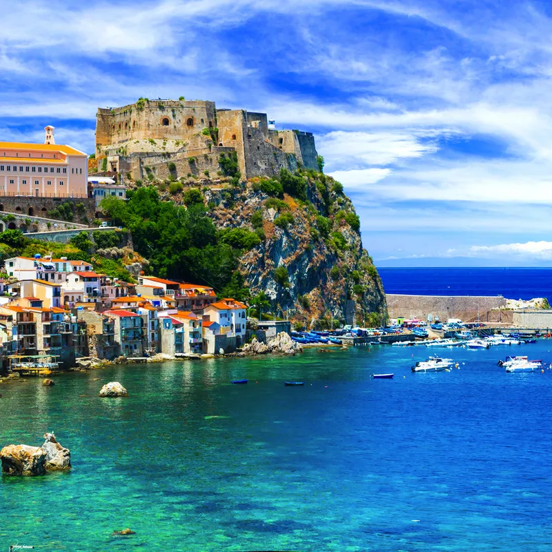 Küstenstadt mit farbenfrohen Gebäuden und einer Burg auf einem Hügel am Meer.