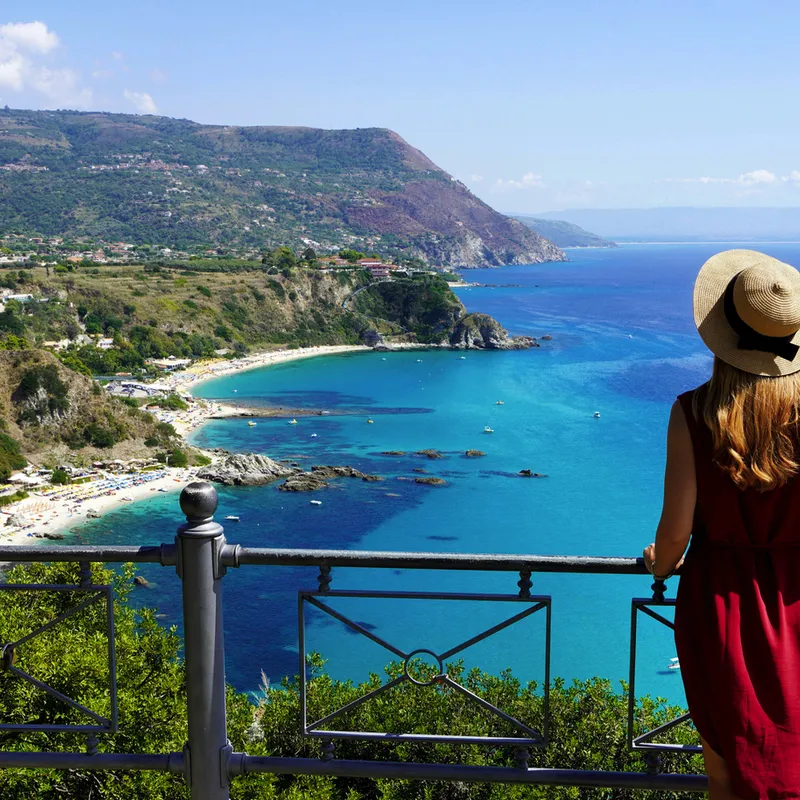 Frau mit Hut blickt von Balkon auf Küste mit blauem Meer und grüner Landschaft.