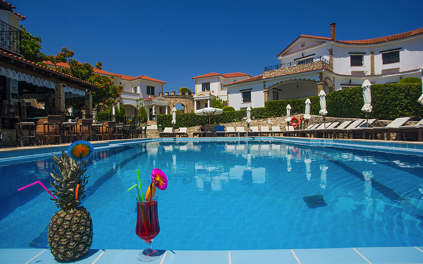 Hotelpool mit Getränken und Liegestühlen, umgeben von weißen Gebäuden.