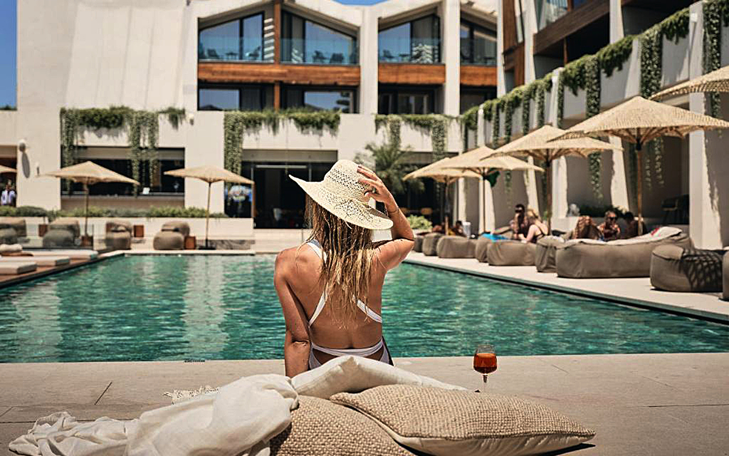 Frau im Bikini sitzt am Pool, umgeben von Sonnenschirmen und modernen Gebäuden