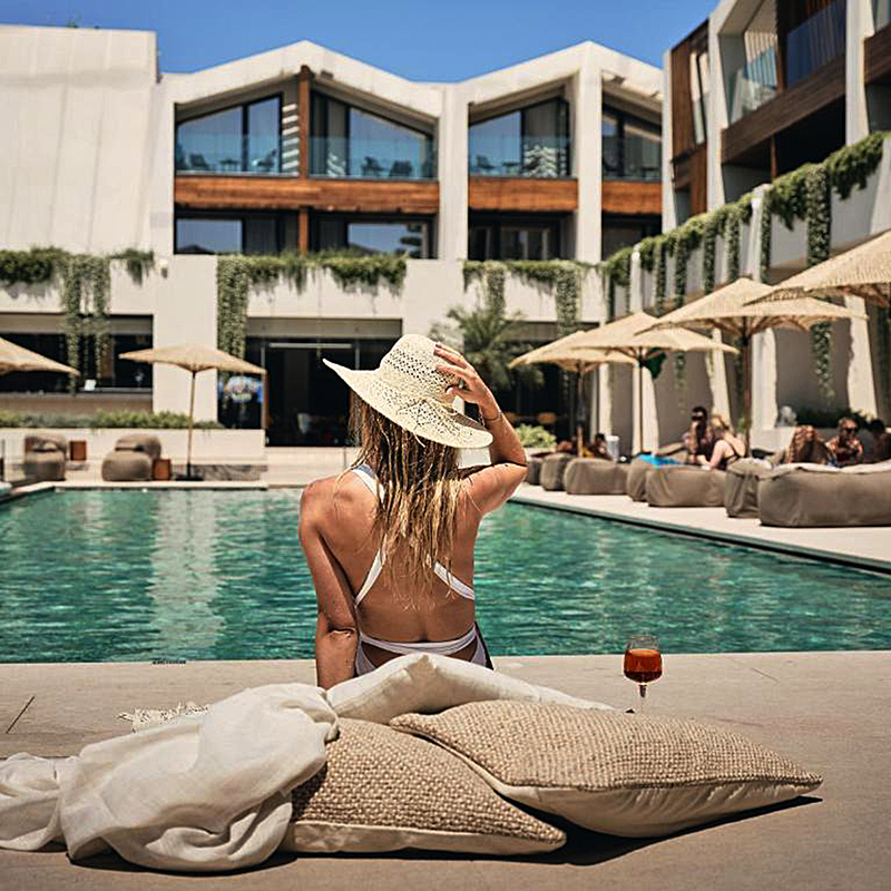 Frau im Bikini sitzt am Pool, umgeben von Sonnenschirmen und modernen Gebäuden