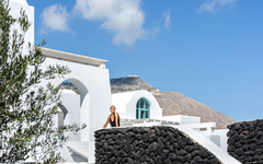 Frau auf Hotelterrasse vor weißer Architektur und Bergen in Santorin.