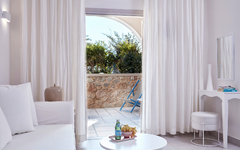 Helles Hotelzimmer mit Blick auf eine Terrasse und Garten, weißes Sofa und Tisch mit Wasserflasche.