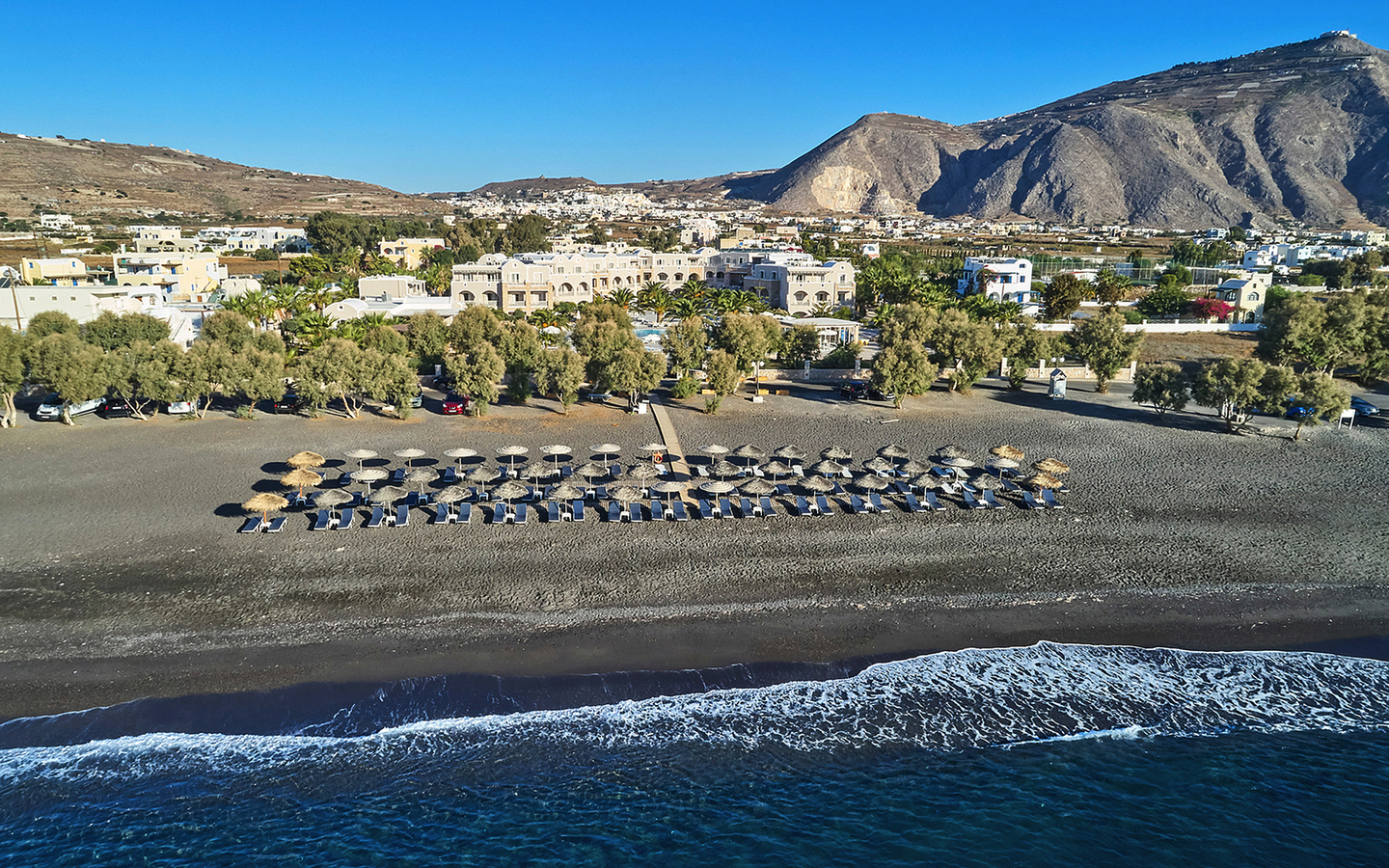 Strand mit Liegestühlen und Sonnenschirmen vor Hotel und Bergen im Hintergrund.
