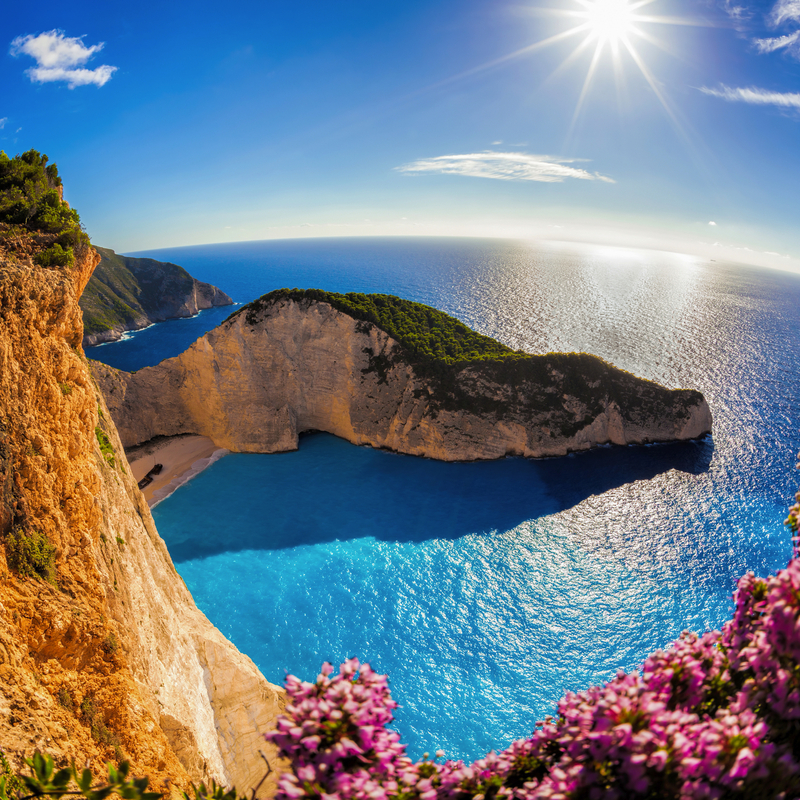 Steilküste mit blauem Meer, Strand und blühenden Pflanzen unter sonnigem Himmel.