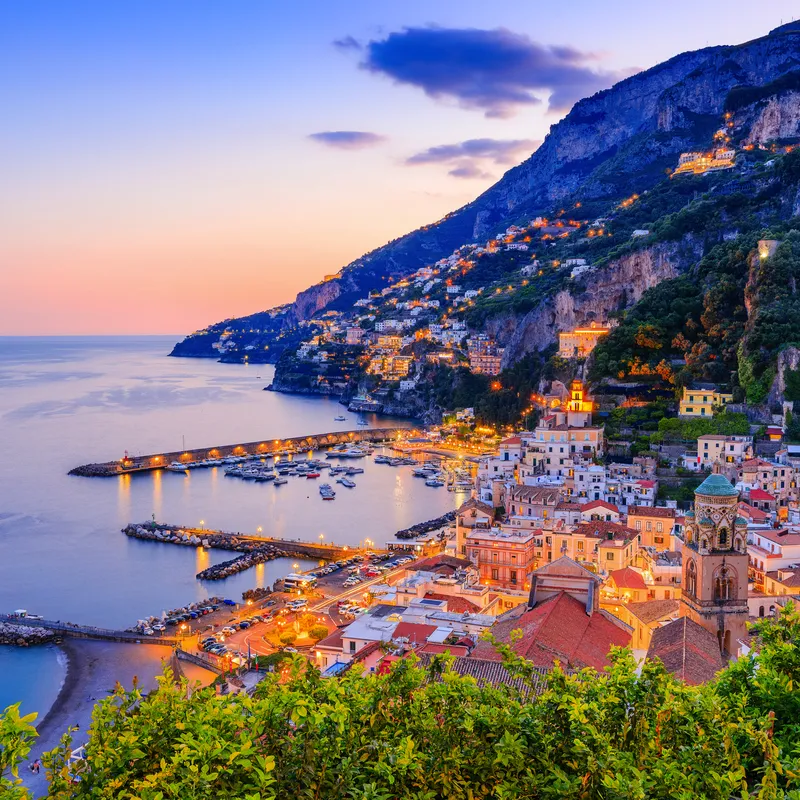 Küstenstadt bei Sonnenuntergang mit beleuchteten Häusern und Hafen am Meer.