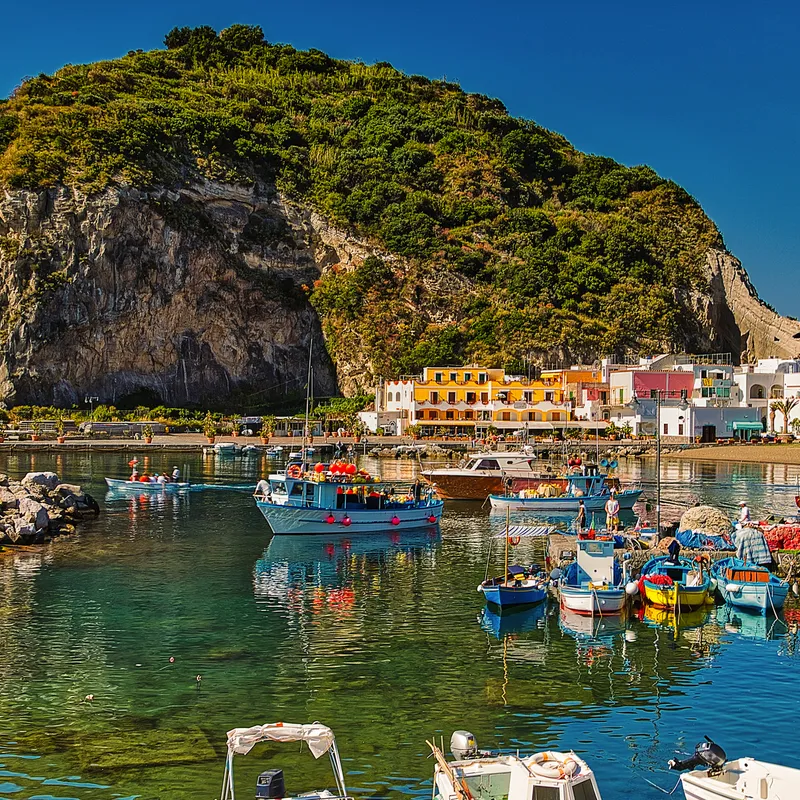 Hafen mit bunten Booten vor einem bewachsenen Felsen und bunten Gebäuden.