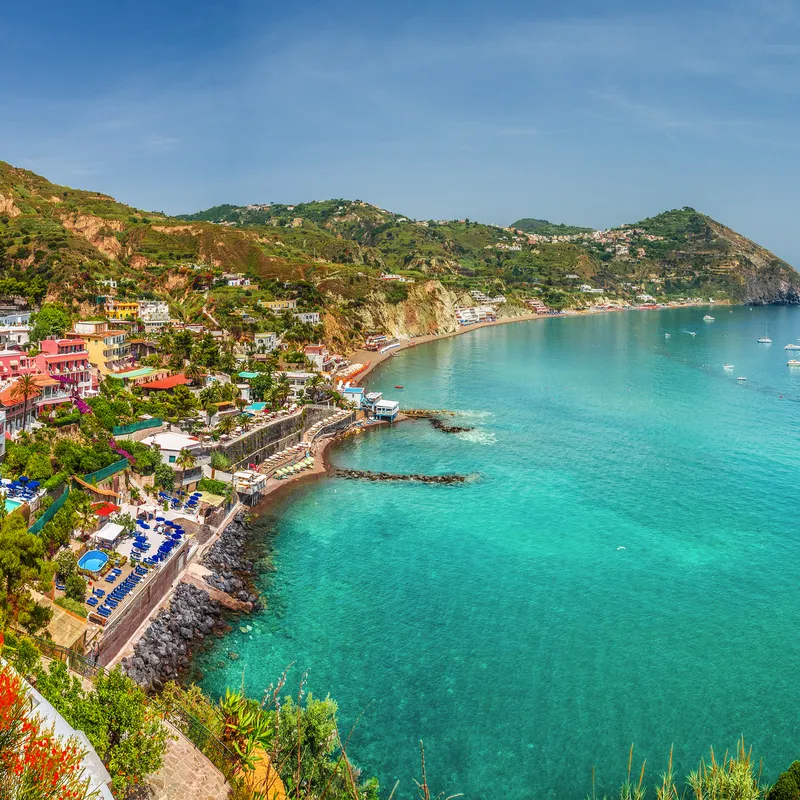Küstenstadt mit bunten Häusern am türkisblauen Meer und grünen Hügeln.