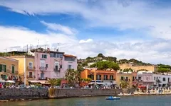 Häuser entlang einer Uferpromenade bei sonnigem Wetter und blauem Himmel.