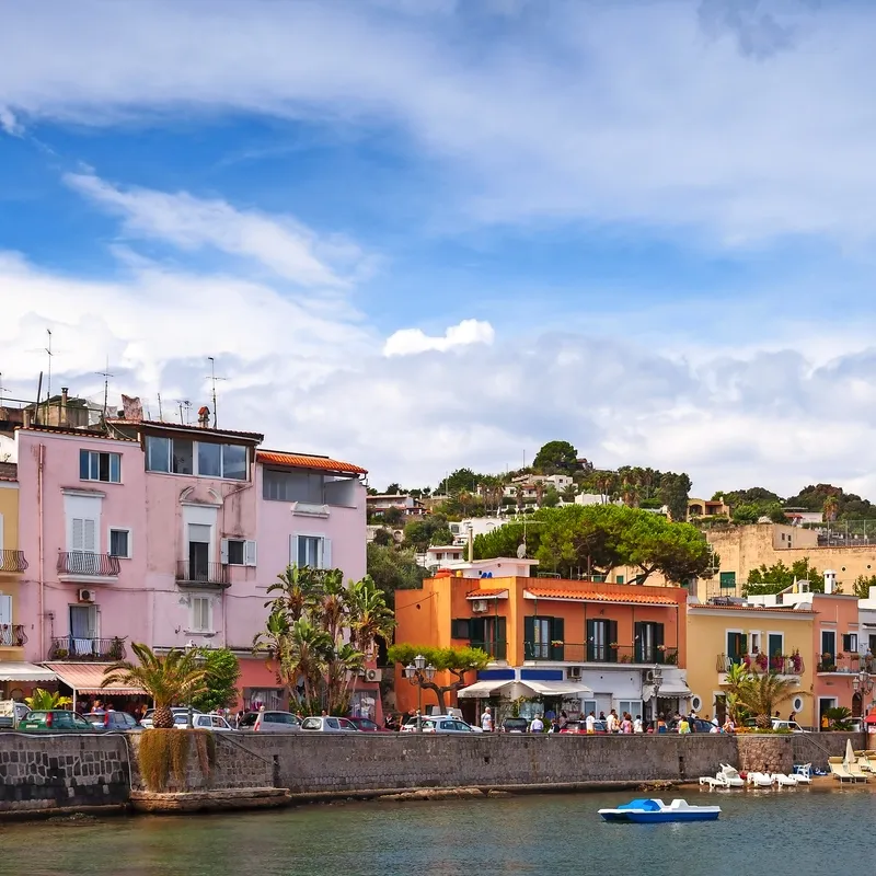 Häuser entlang einer Uferpromenade bei sonnigem Wetter und blauem Himmel.