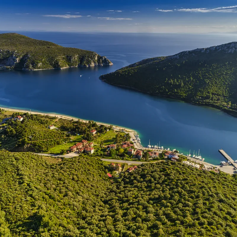 Bucht mit blauem Wasser und grünen Hügeln, kleine Gebäude und Boote am Ufer.