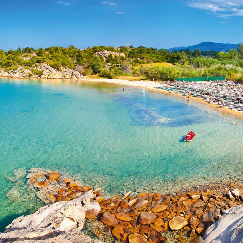Strand mit klarem Wasser, Sonnenschirmen und bewaldeter Küste im Hintergrund.