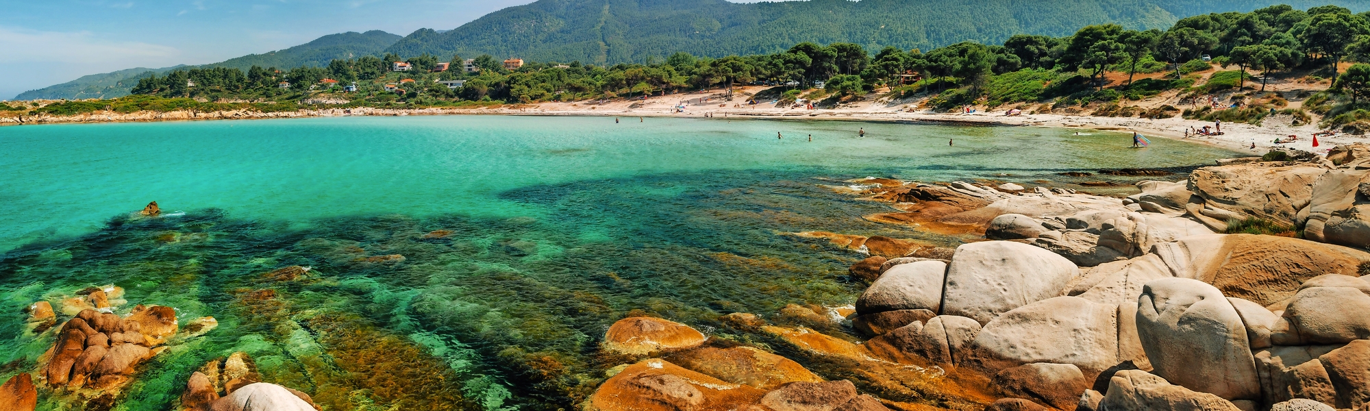 Küstenlandschaft mit klarem Wasser, Felsen und bewaldeten Hügeln im Hintergrund.