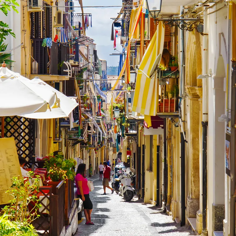 Belebte enge Straße in mediterraner Stadt mit Balkonen und Markisen.