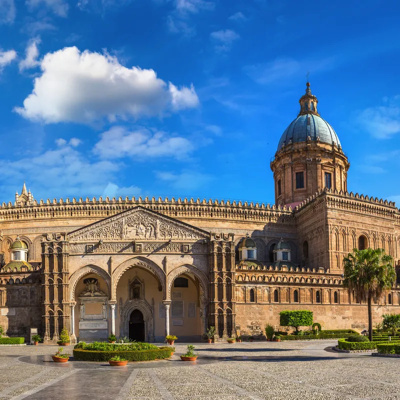 Kathedrale von Palermo an einem sonnigen Tag, umgeben von Palmen und historischer Architektur.