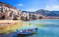 Boot auf klarem Wasser vor Küste mit Häusern und Bergen im Hintergrund bei Sonnenschein