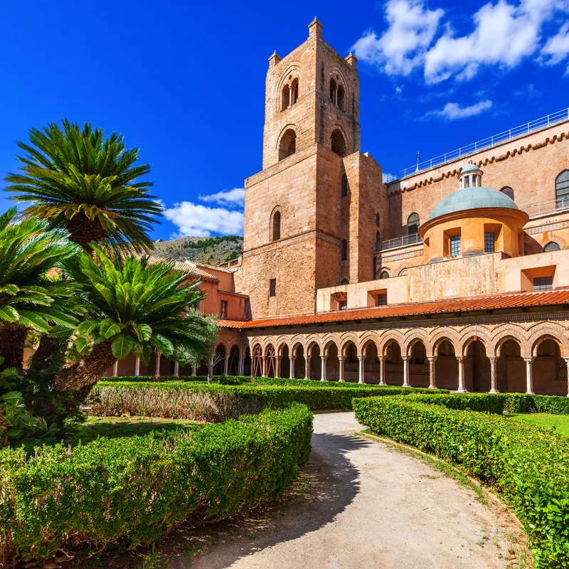 Kreuzgang eines Klosters mit Palmen und historischem Turm unter blauem Himmel.