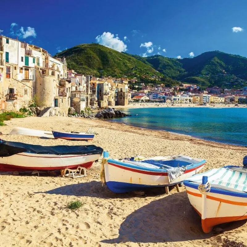 Boote liegen am Strand in einer mediterranen Küstenstadt vor grünen Hügeln und blauem Meer.