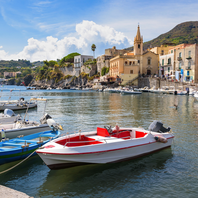Boote in einem kleinen Hafen vor bunten Häusern und einem Kirchgebäude, Hügel im Hintergrund.