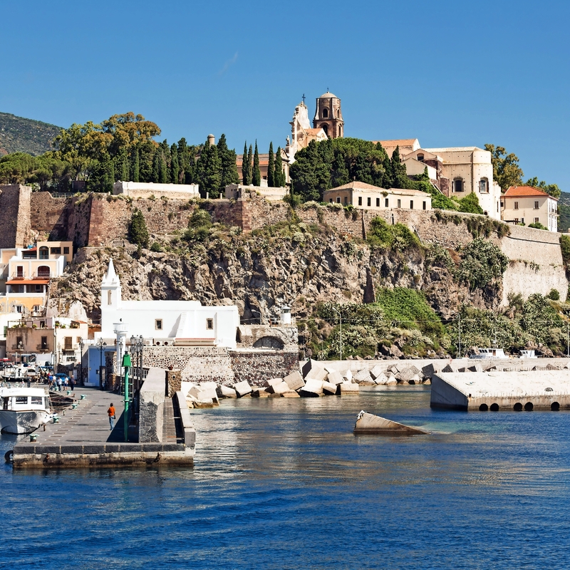 Küstenstadt mit Kirche auf einem Hügel, Boote im Hafen, sonnige Landschaft.