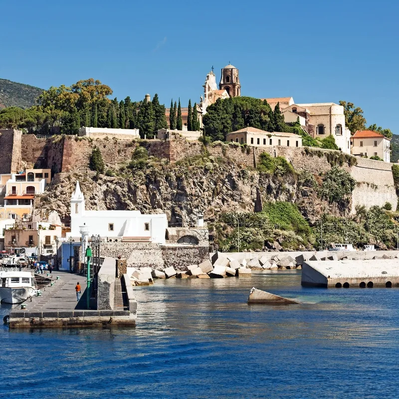 Küstenstadt mit Kirche auf einem Hügel, Boote im Hafen, sonnige Landschaft.