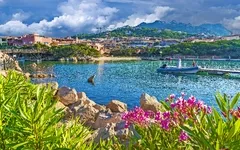 Küstenlandschaft mit Booten, bunten Blumen und Gebäuden vor einer Bergkulisse.