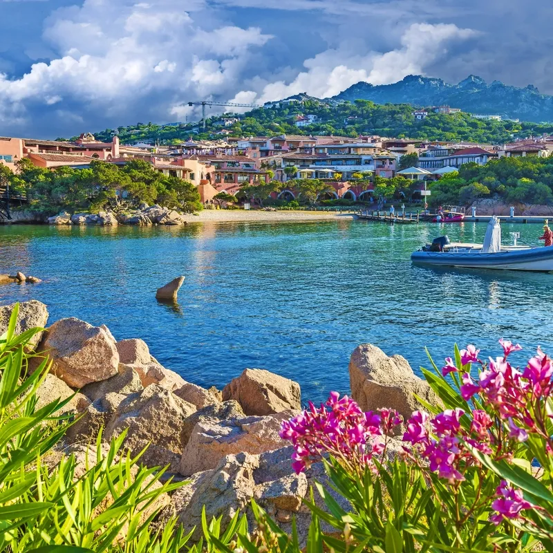 Küstenlandschaft mit Booten, bunten Blumen und Gebäuden vor einer Bergkulisse.