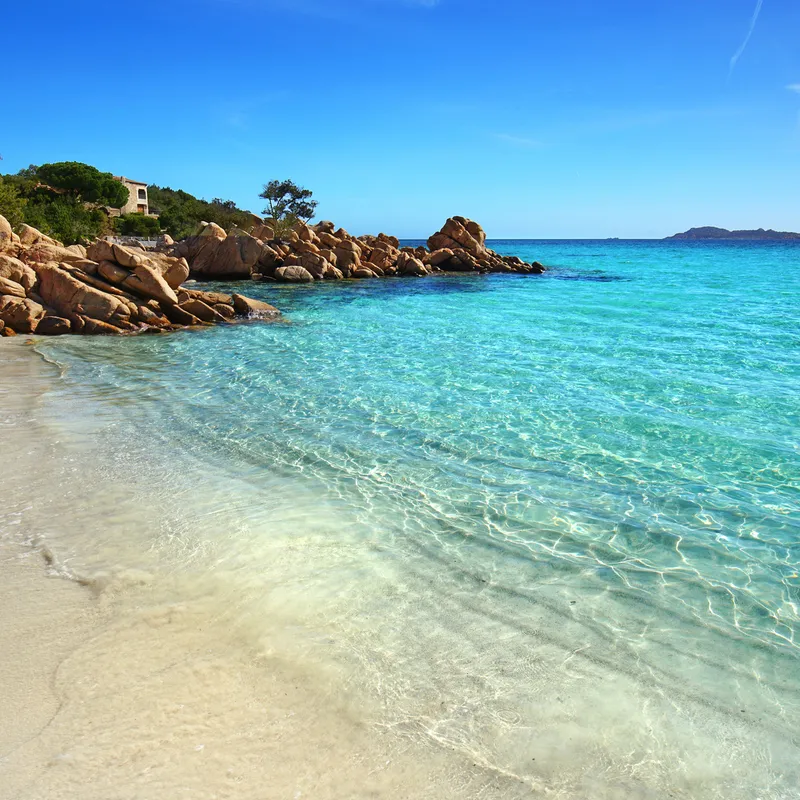 Sandstrand mit klarem türkisfarbenem Meer und Felsen unter blauem Himmel.