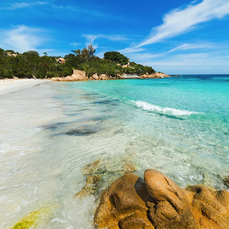 Klares Wasser und Sandstrand mit Felsen und Bäumen im Hintergrund.