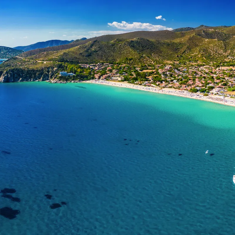 Küste mit türkisfarbenem Wasser und Booten, Hügel im Hintergrund.