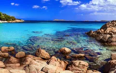 Klares, türkisfarbenes Meereswasser mit Felsen und Bäumen unter blauem Himmel.