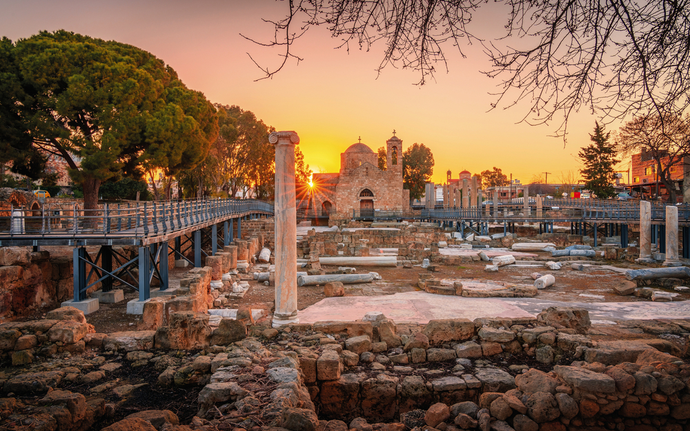 Archäologische Stätte in Paphos, Zypern, bei Sonnenuntergang mit alter Kirche im Hintergrund.