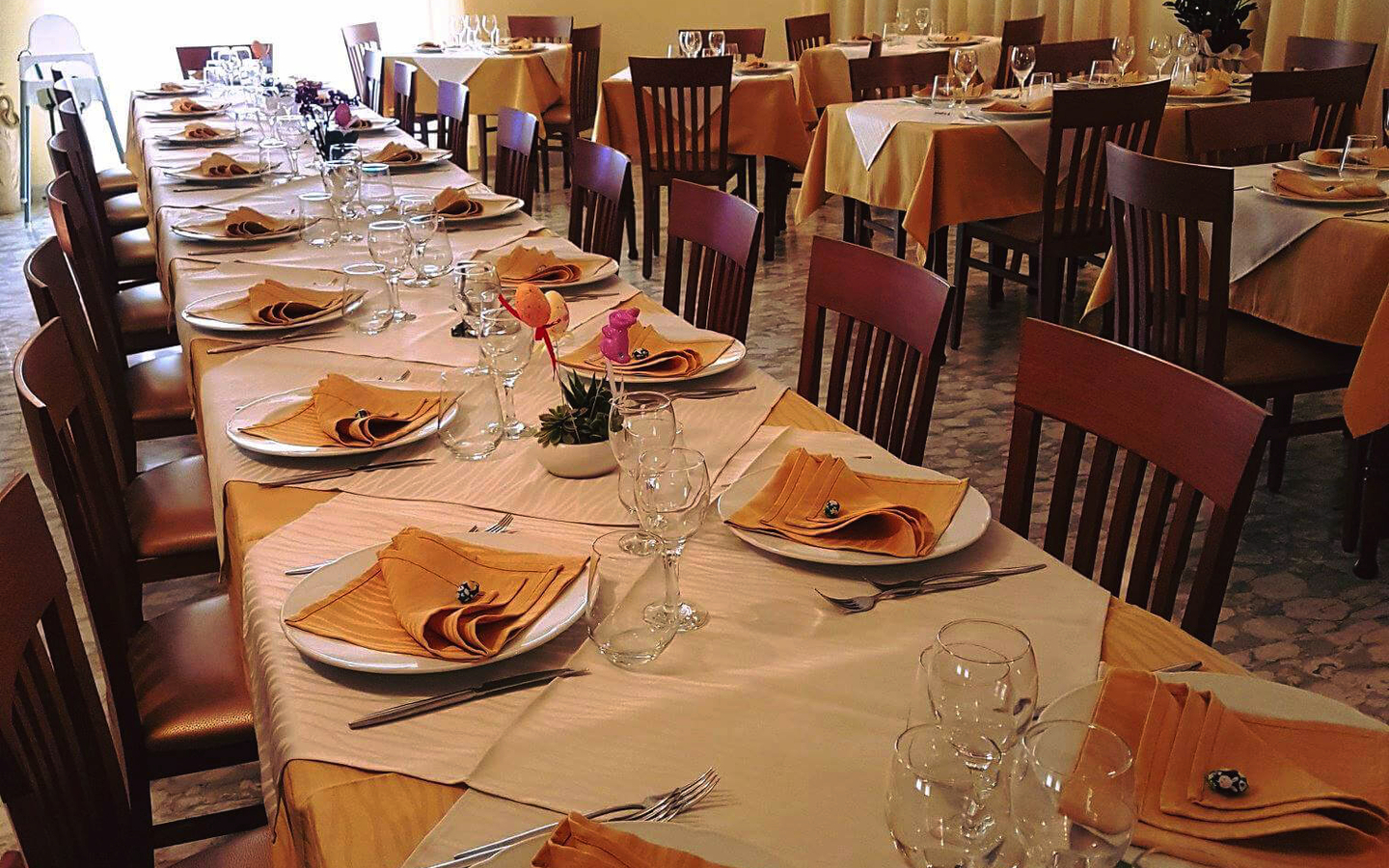 Restaurant mit gedeckten Tischen, gelbe Servietten, Holzstühle, helle Vorhänge.