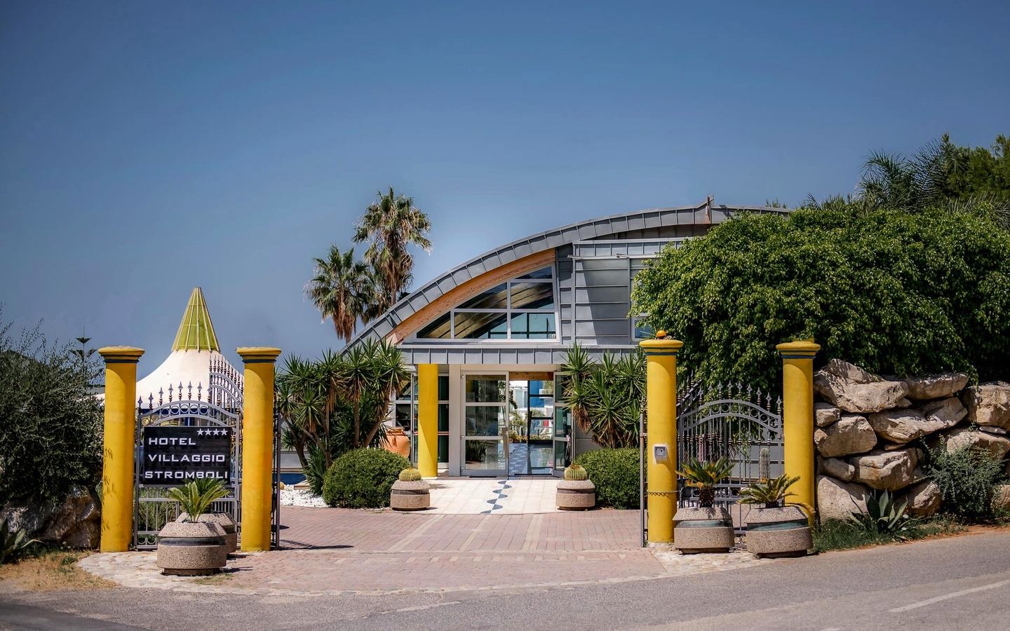 Eingang des Hotel Villaggio Stromboli in Kalabrien, umgeben von Palmen und Pflanzen.
