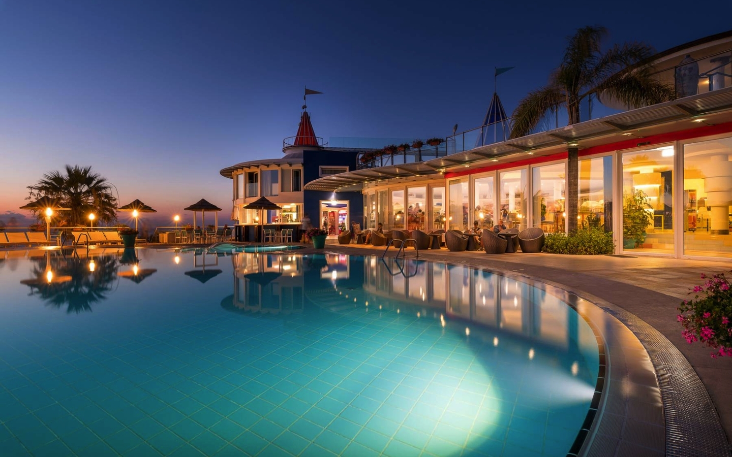 Hotel mit beleuchtetem Pool bei Abenddämmerung und Palmen in Kalabrien, Italien.