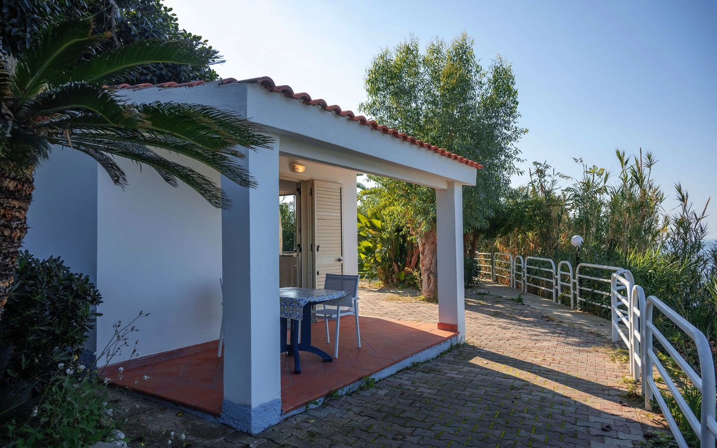 Kleines Bungalow mit Terrasse und Gartenmöbeln, umgeben von Pflanzen und Bäumen.
