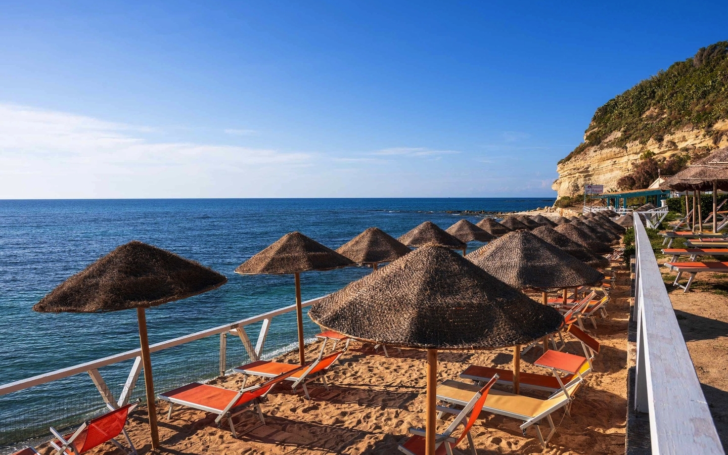 Sonnenschirme und Liegestühle am Sandstrand mit Meerblick bei klarem Himmel.