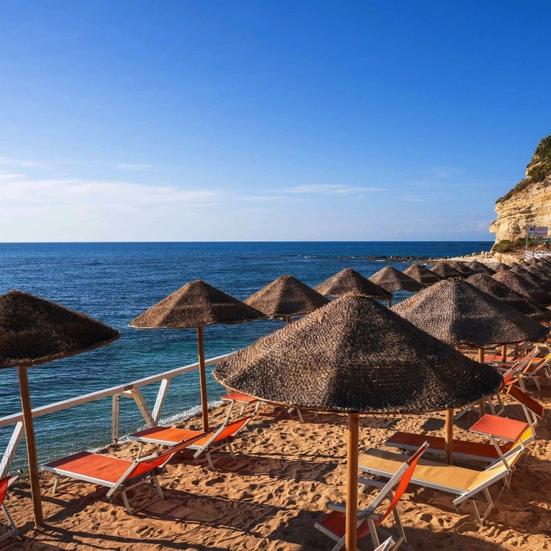 Sonnenschirme und Liegestühle am Sandstrand mit Meerblick bei klarem Himmel.