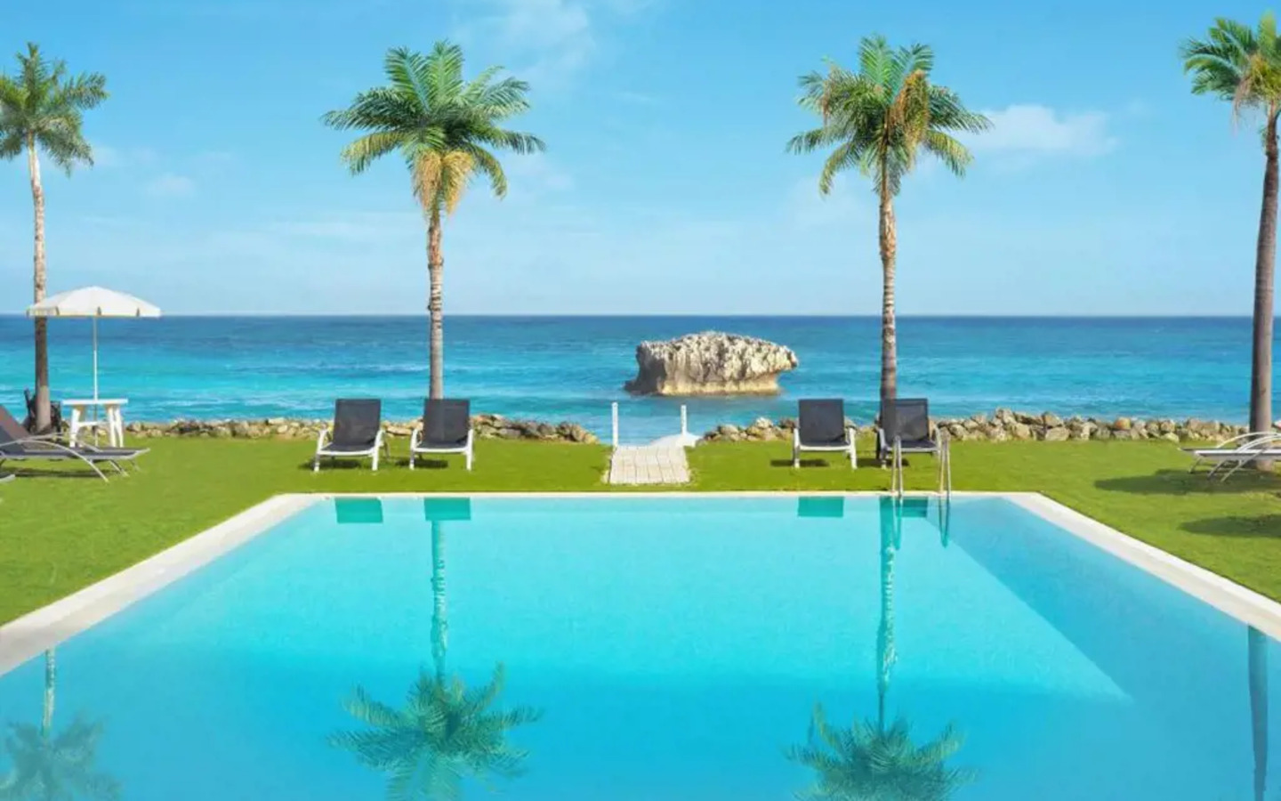 Pool mit Liegestühlen und Palmen, im Hintergrund das Meer bei klarer Himmel.