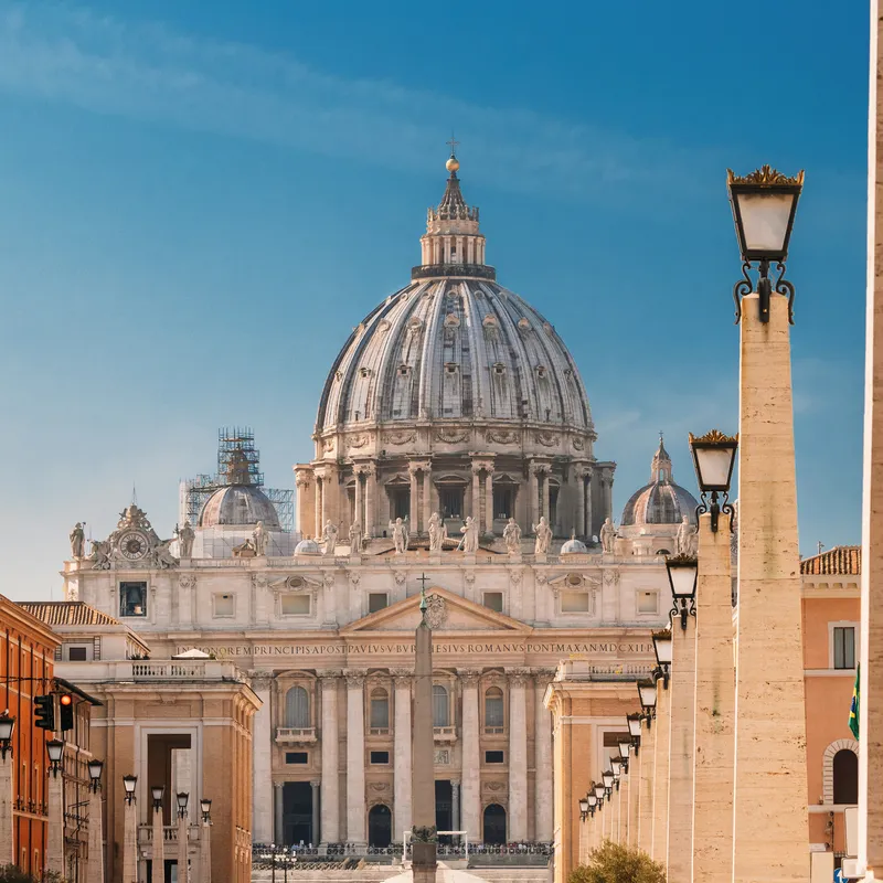 Kuppel des Petersdoms in Rom, umgeben von Säulen und blauen Himmel.