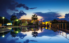 Hotelpool bei Sonnenuntergang mit Spiegelung im Wasser, Palmen und beleuchtetem Himmel.