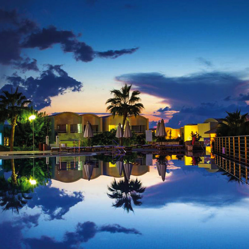 Hotelpool bei Sonnenuntergang mit Spiegelung im Wasser, Palmen und beleuchtetem Himmel.