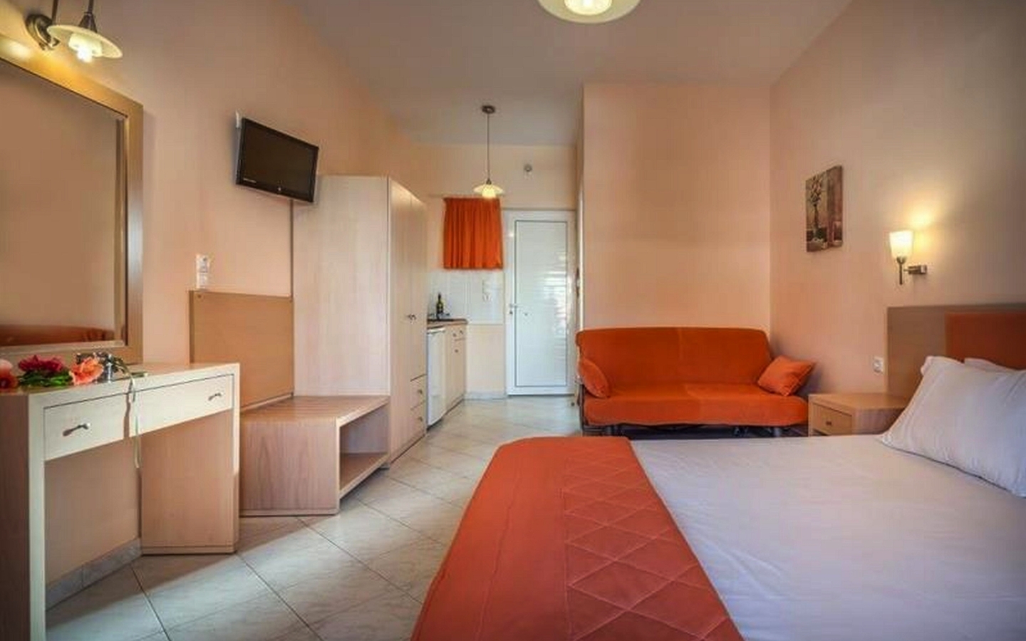 Modernes Hotelzimmer mit Bett, Sofa und kleinem Küchenbereich in warmen Farbtönen.