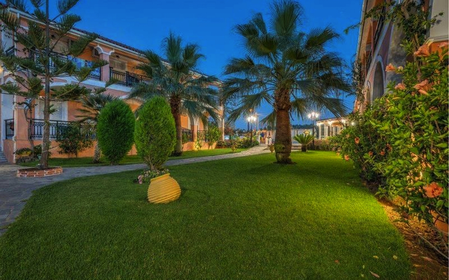 Gartenansicht eines Hotels mit Palmen und beleuchtetem Gehweg bei Dämmerung.