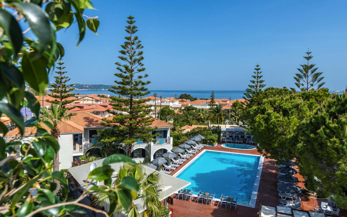 Blick auf einen Hotelpool mit Sonnenschirmen und Bäumen, im Hintergrund das Meer