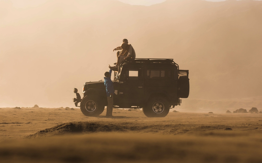 Jeep in sandiger Landschaft mit Personen auf dem Dach und daneben stehend.